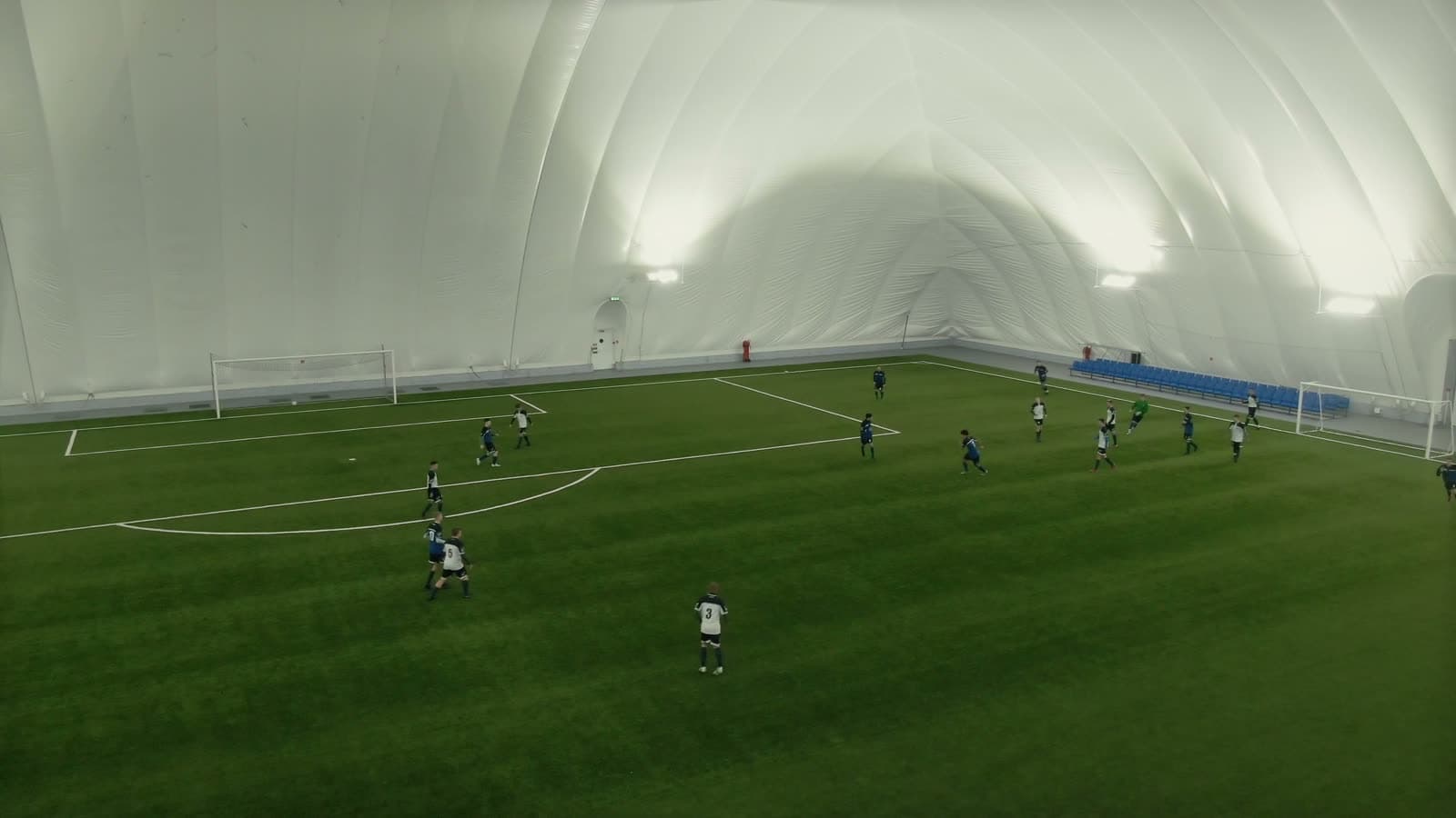 Full view of an indoor soccer dome interior showing the pitch, sideline seating, and match in progress