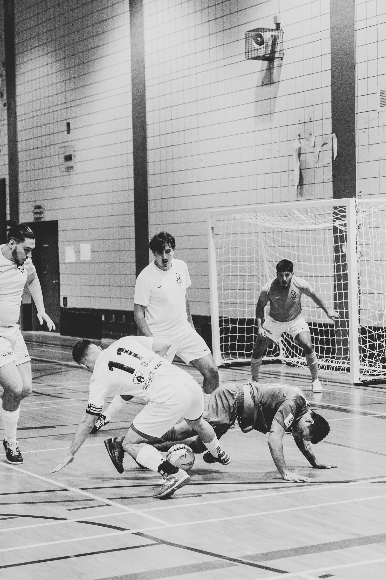 Indoor futsal players in action on a hardcourt facility in black and white, with a goal visible in the background