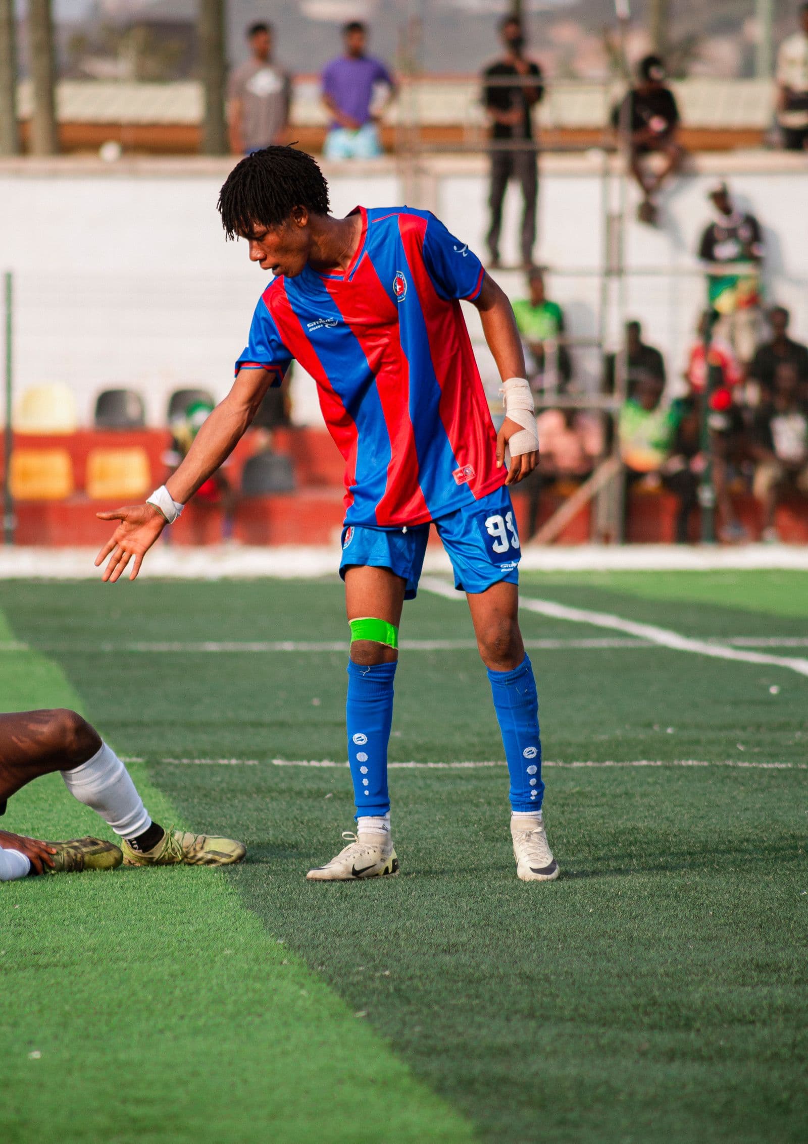 Soccer player in red and blue kit standing on artificial turf during a match with other players visible in the background